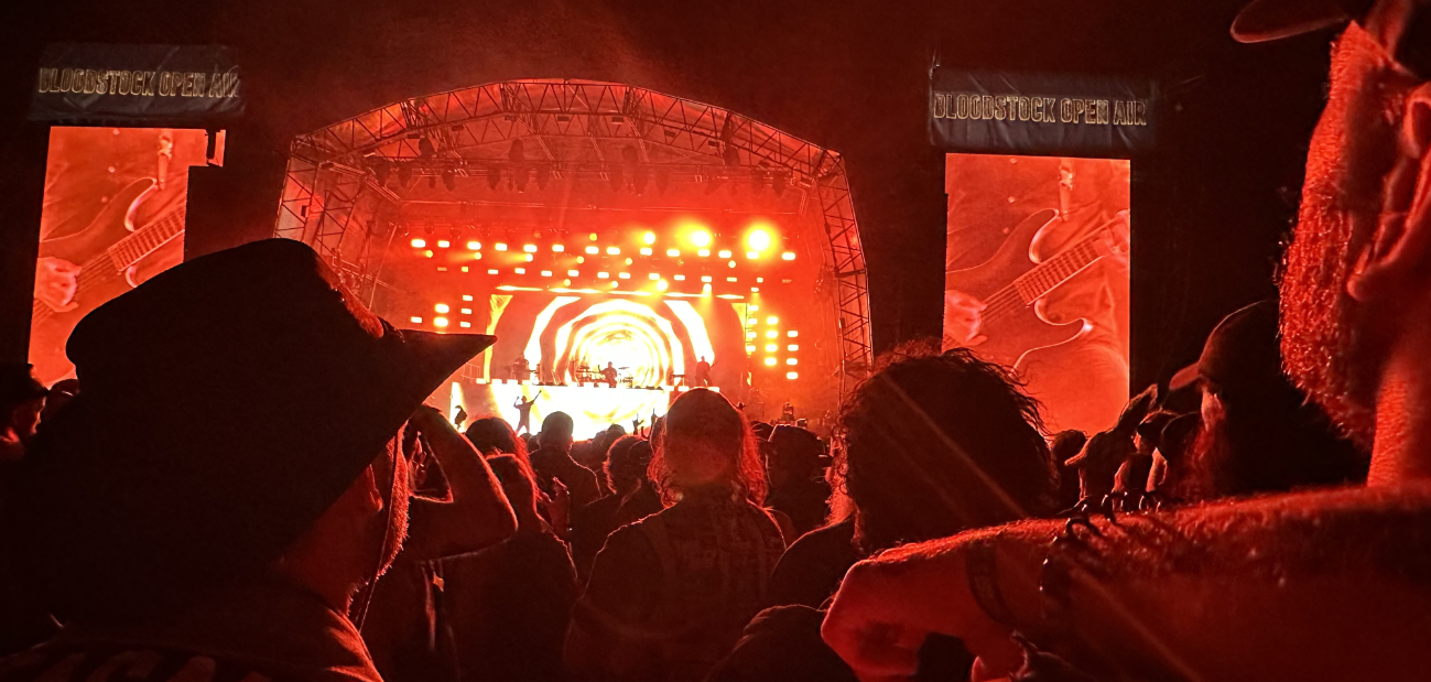 A stage crowd watches a band play. The stage is light in bright red colours. There are two screen on each side showing a close up of the guitar